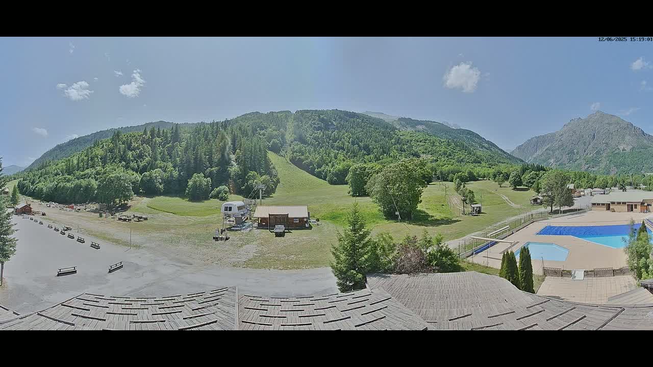 A panoramic view of a mountainous region on a sunny day shows a grassy area with a ski lift, a small building, and an outdoor swimming pool, all nestled among green hills.