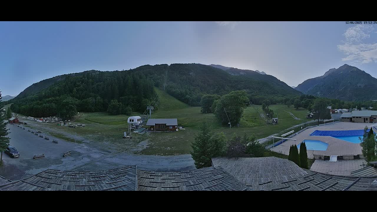 A panoramic view of a mountain valley at dusk, showing a ski lift, a small building, a parking lot, an outdoor swimming pool, and a roof in the foreground.