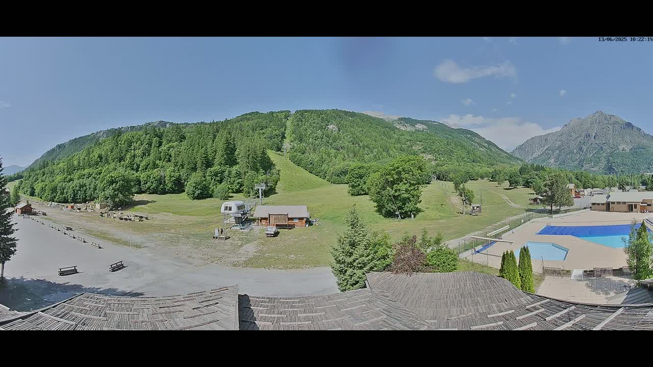 A panoramic view of a mountain resort on a sunny day shows a ski lift, small buildings, a parking lot, and an outdoor swimming pool nestled among green hills and mountains.