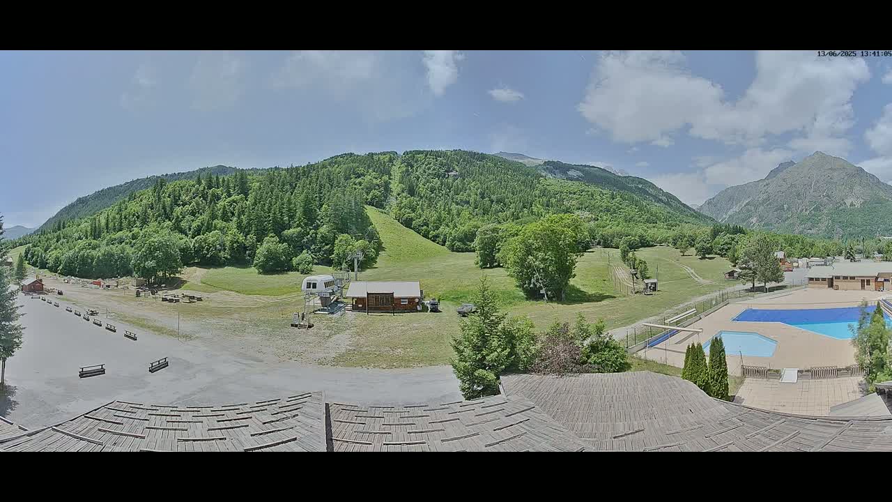 A mostly sunny day shows a mountain valley scene with a ski lift, small buildings, a swimming pool, and a large grassy area.