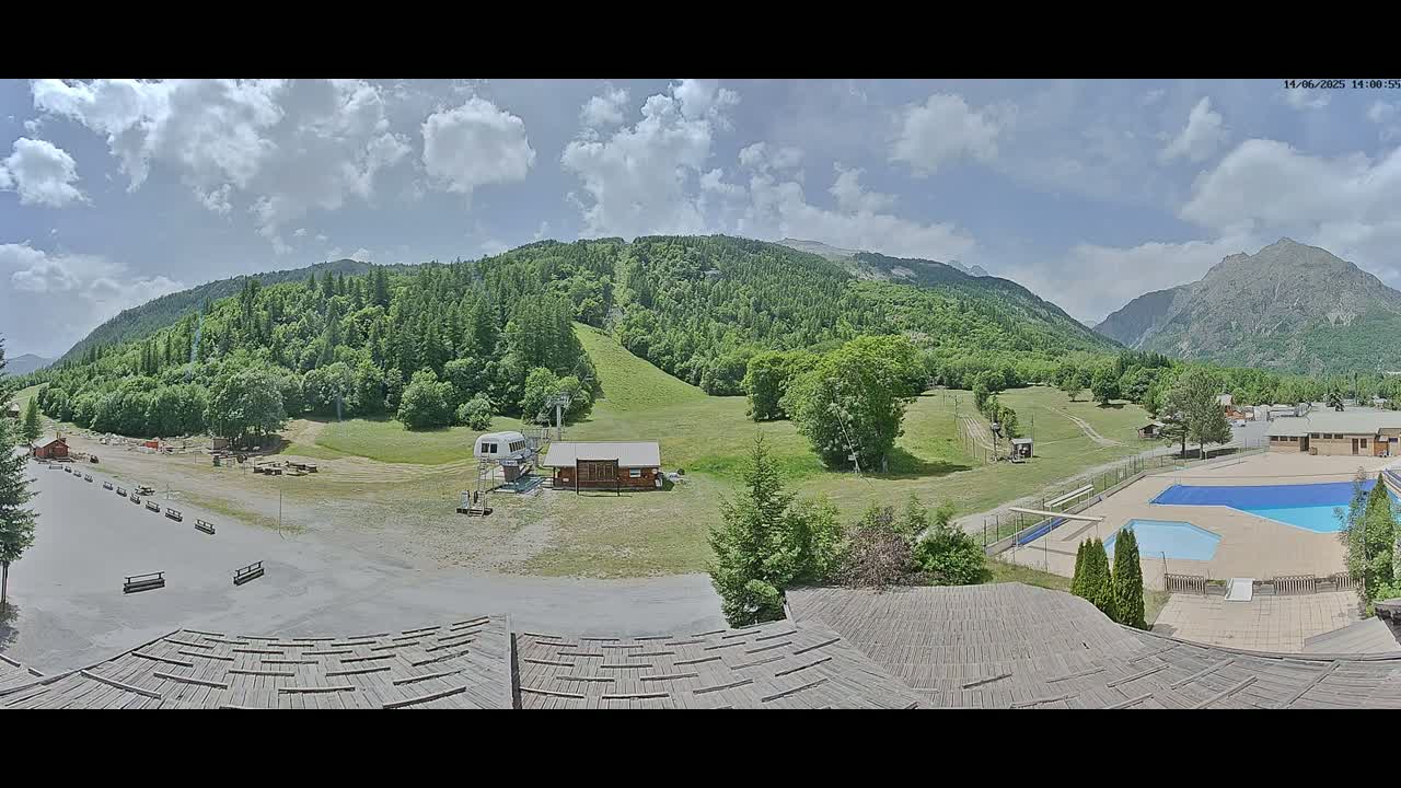 A partly cloudy day reveals a mountain valley scene with a ski lift, a small building, a swimming pool, and a large grassy area.