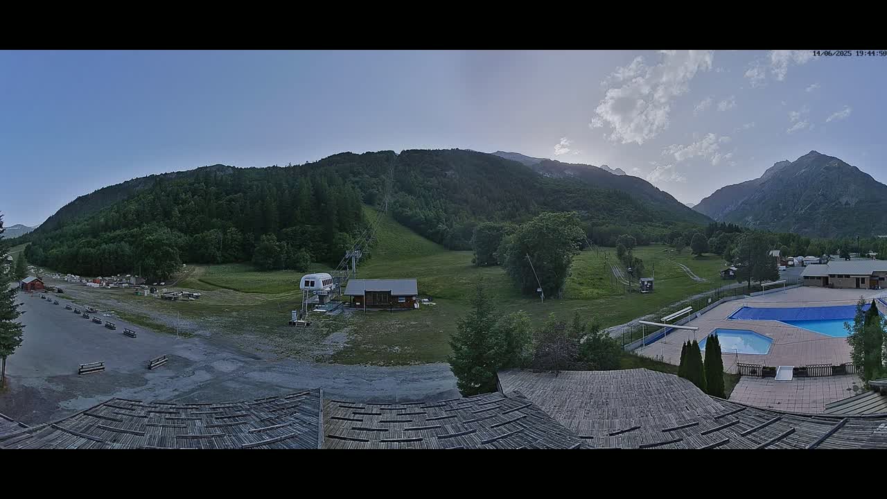 A panoramic view of a mountain valley shows a ski lift, a small building, a swimming pool, and a grassy area under a partly cloudy sky.