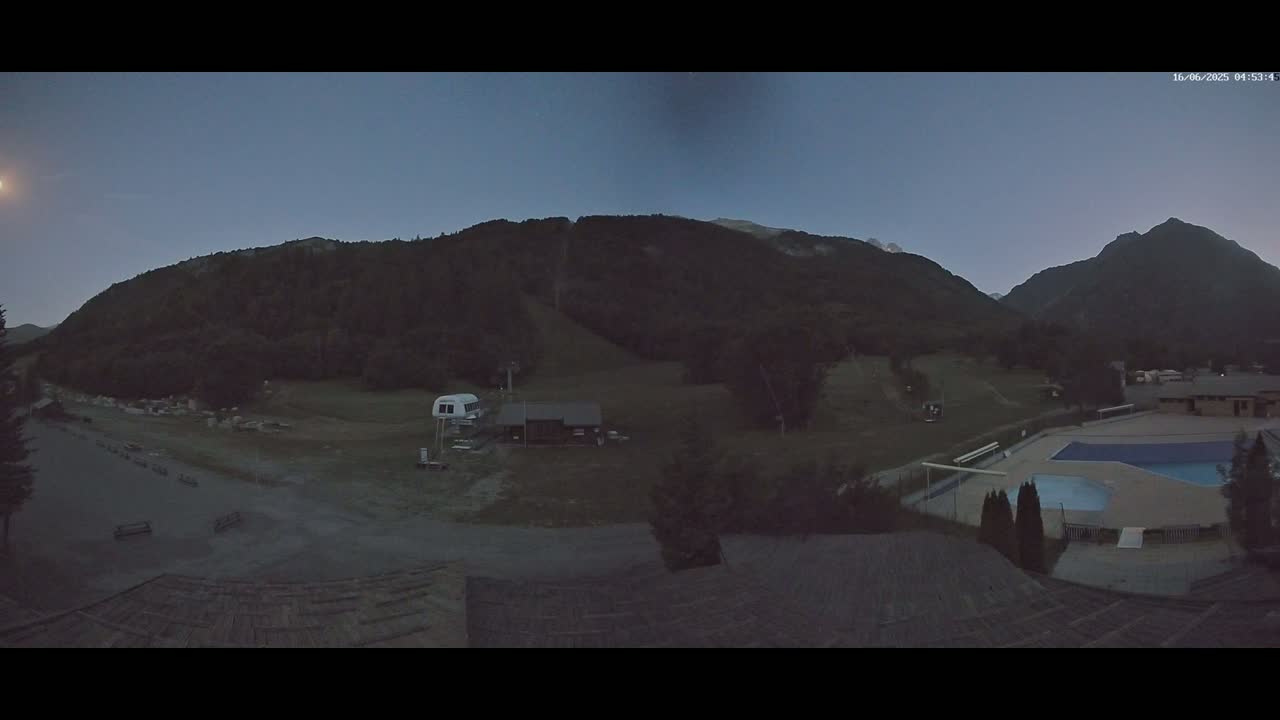 A nighttime view shows a ski lift, a small building, and an outdoor swimming pool nestled in a valley between dark, tree-covered mountains under a clear, twilight sky.
