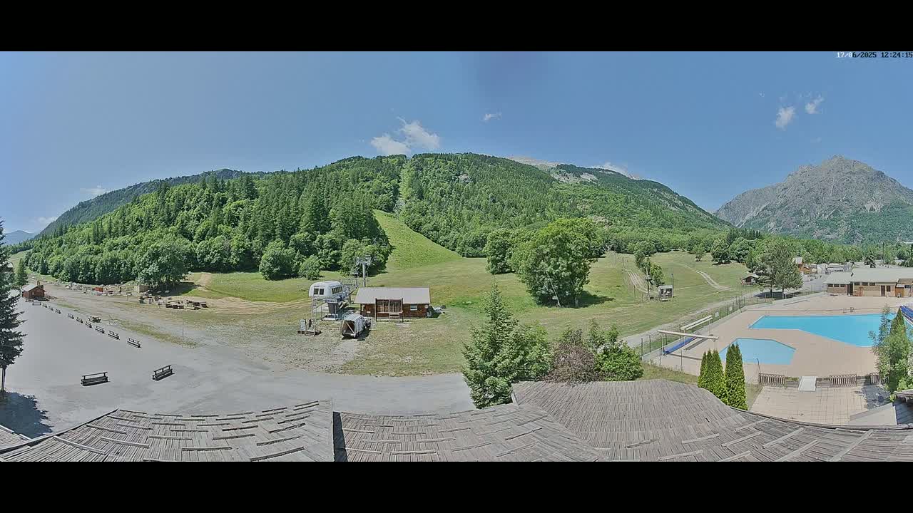 A panoramic view of a mountainside resort on a sunny day, showing a grassy area with a chairlift, buildings, and an outdoor swimming pool.