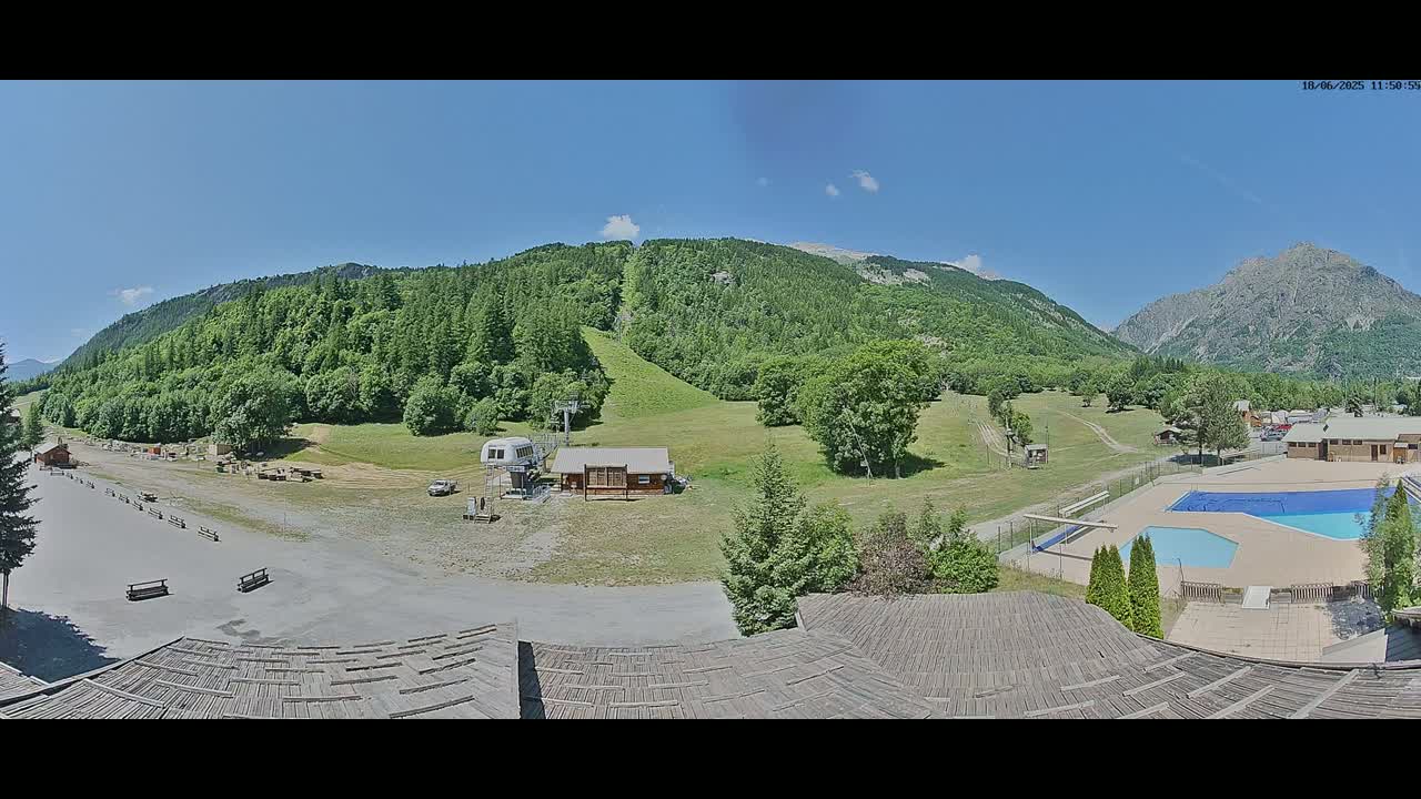 A panoramic view on a sunny day shows a mountain resort with a grassy ski slope, a small building, a swimming pool, and a large parking area.