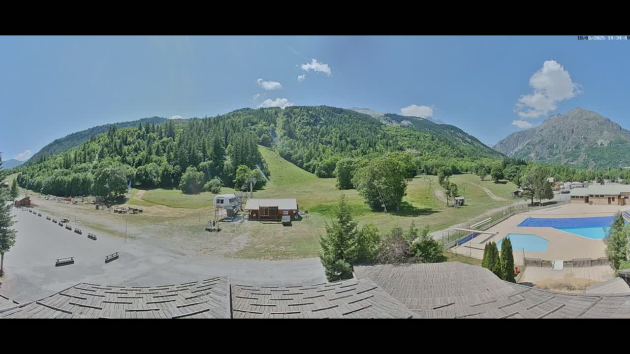 A sunny day reveals a mountain valley scene with green hills, a ski lift, a small building, a swimming pool, and a partially visible building in the foreground.