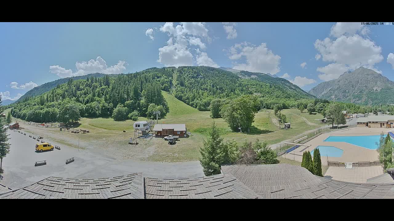 A mountain valley scene under a partly cloudy sky shows a parking lot, a small building, a chairlift, a swimming pool, and several other structures nestled among green hills and mountains.