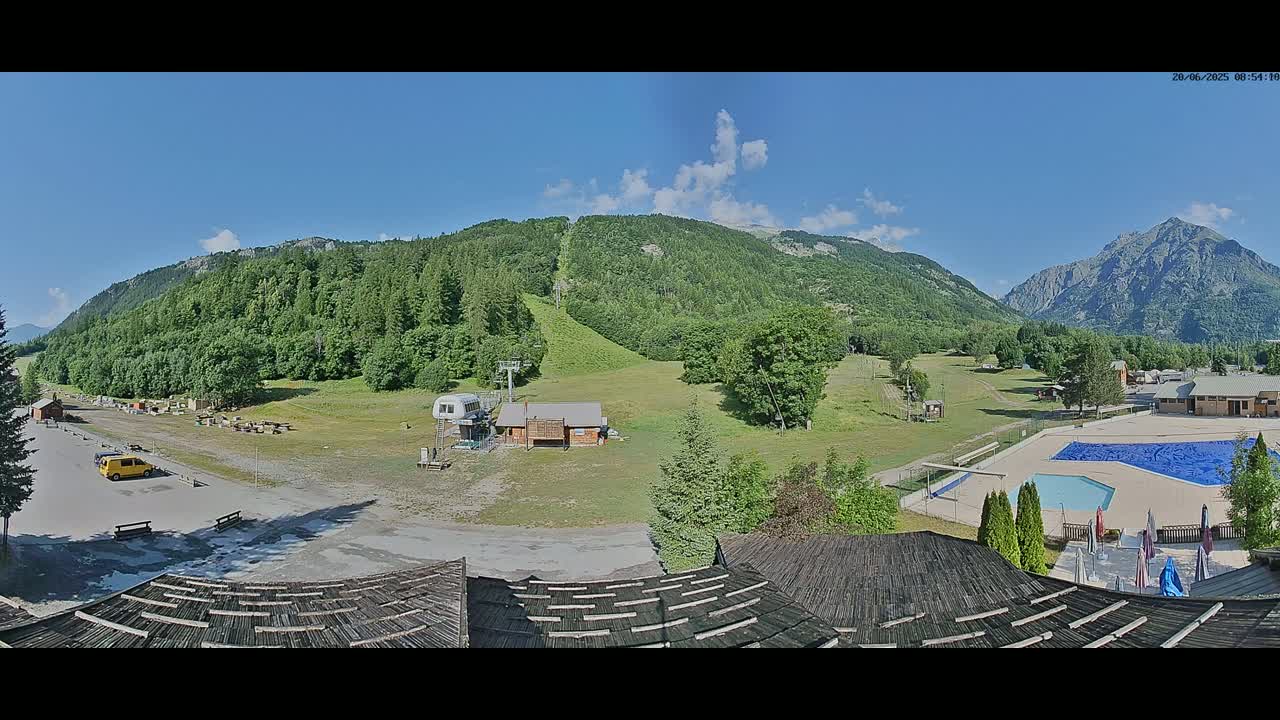 A mountain valley scene on a sunny day shows a ski lift, a small building, a parking lot, a swimming pool, and a large forested mountain in the background.