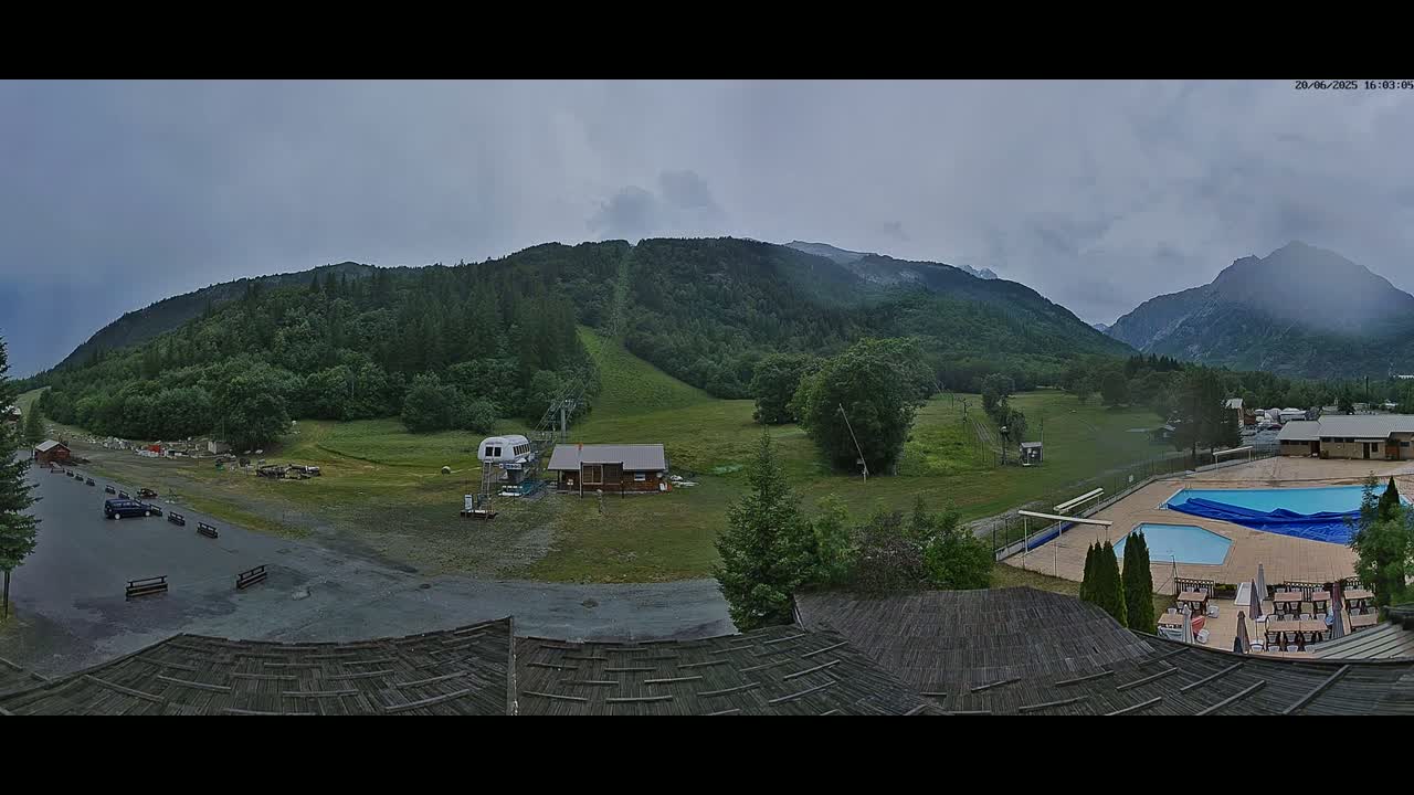 A mountain valley scene under an overcast sky shows a ski lift, small buildings, a swimming pool, and a parking lot, with mountains in the background.