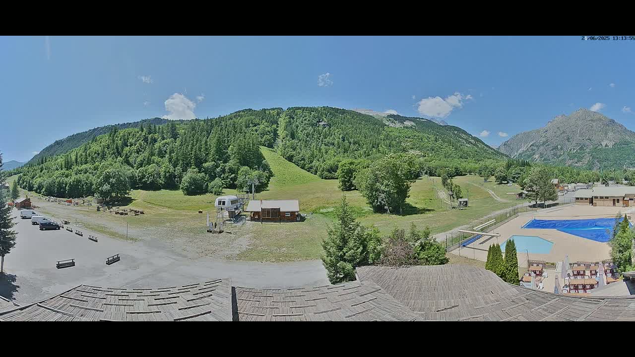 A sunny mountain valley scene shows a grassy area with a ski lift, buildings, a swimming pool, and a parking lot, all surrounded by lush green trees and mountains under a mostly clear blue sky.