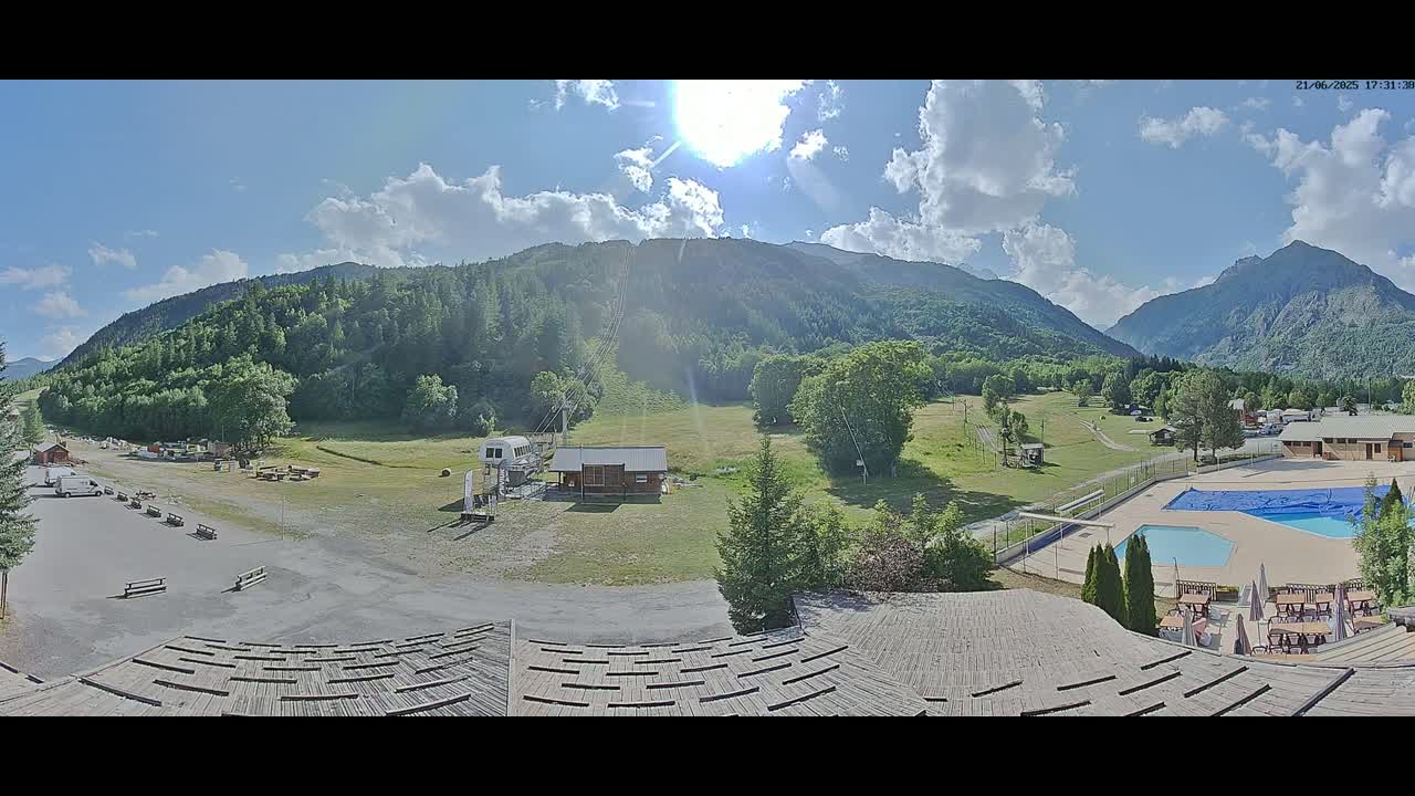 A sunny day reveals a mountain valley scene with a grassy field, a chairlift, a small building, a swimming pool, and outdoor seating areas, all nestled against a backdrop of lush green mountains under a partly cloudy sky.