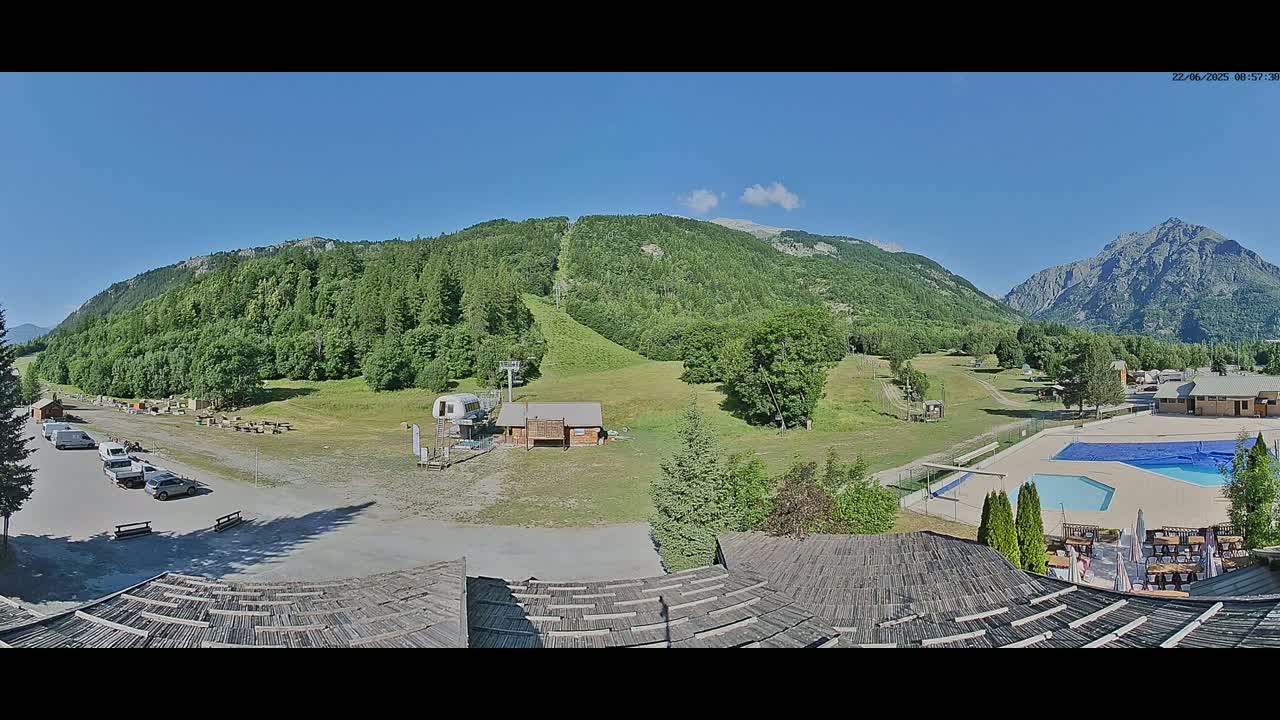 A mountainside view on a sunny day shows a parking lot, a ski lift, a grassy area, a swimming pool, and a building with a thatched roof, all nestled among green hills and mountains.