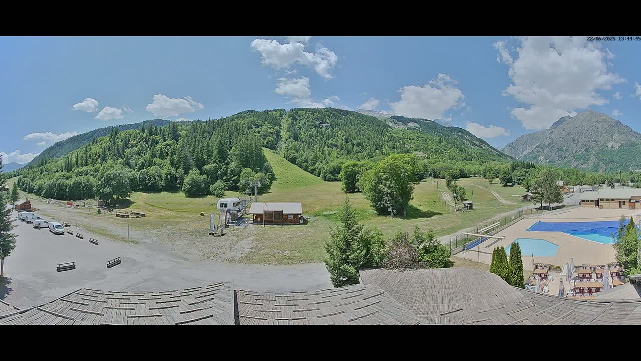 A sunny day reveals a mountainous landscape with a grassy field, a small building, a parking lot with vehicles, a swimming pool, and a wooden structure in the foreground.