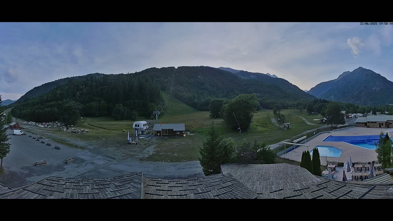 A mountain valley scene under a mostly clear twilight sky shows a chairlift, a small building, a swimming pool, and a parking lot, all situated amongst green fields and forested hills.