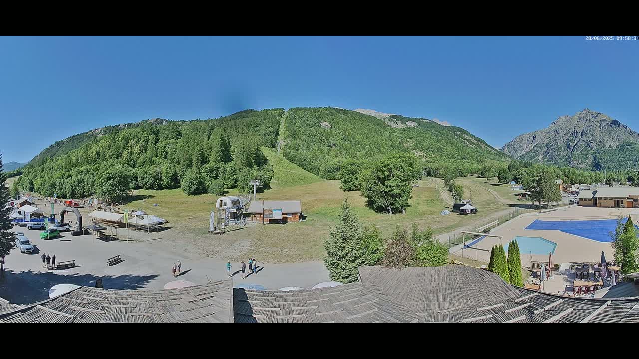 A sunny mountain valley scene shows a parking lot with several people, a grassy area with a ski lift, a swimming pool, and buildings, all set against a backdrop of verdant hills and mountains.