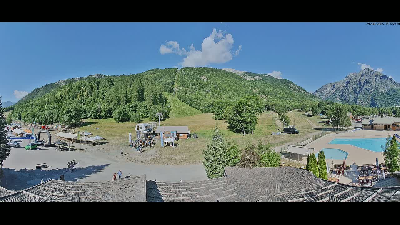 A sunny mountain valley scene shows a parking area, an outdoor pool, a ski lift, and green hillsides under a partly cloudy blue sky.