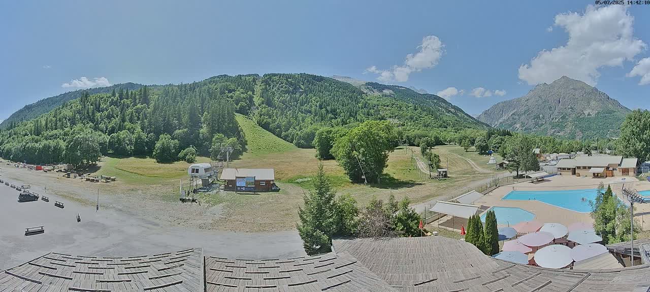 A mountainside view on a sunny day shows a grassy area with a ski lift, a pool, and buildings, all surrounded by green hills and mountains.