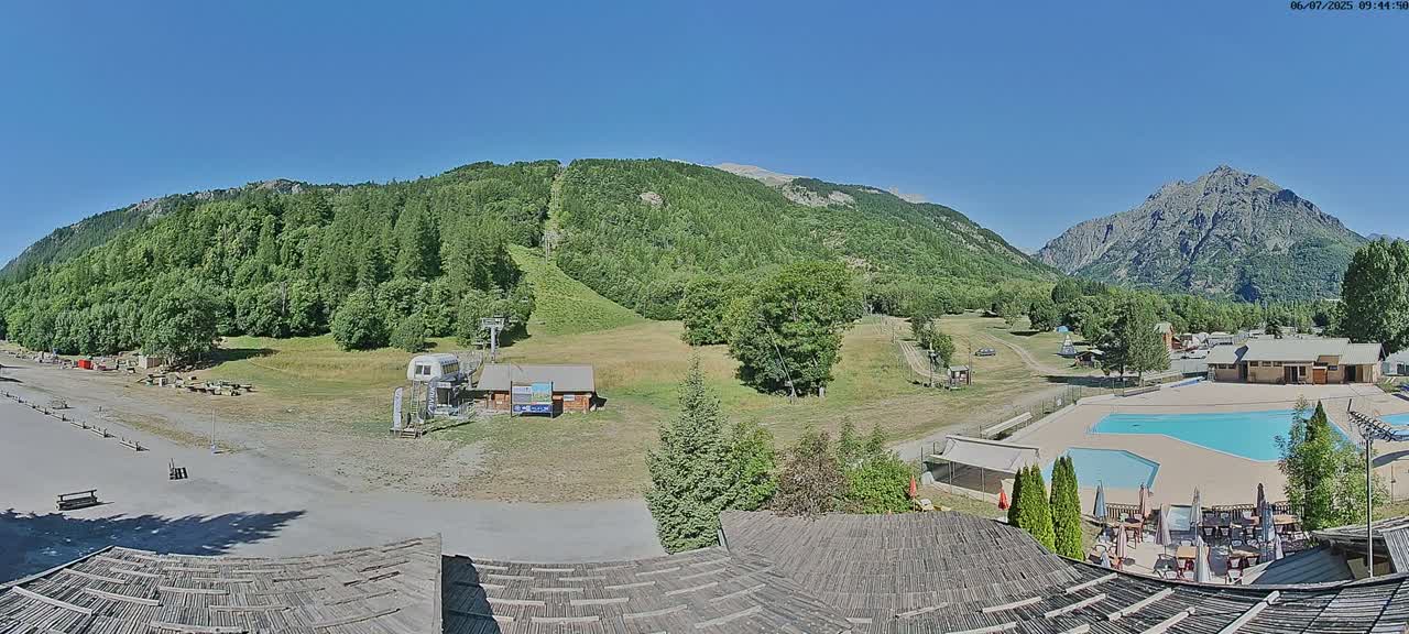 A mountainside view on a sunny day shows a ski lift, a grassy field, a swimming pool, and buildings nestled among green trees and mountains.