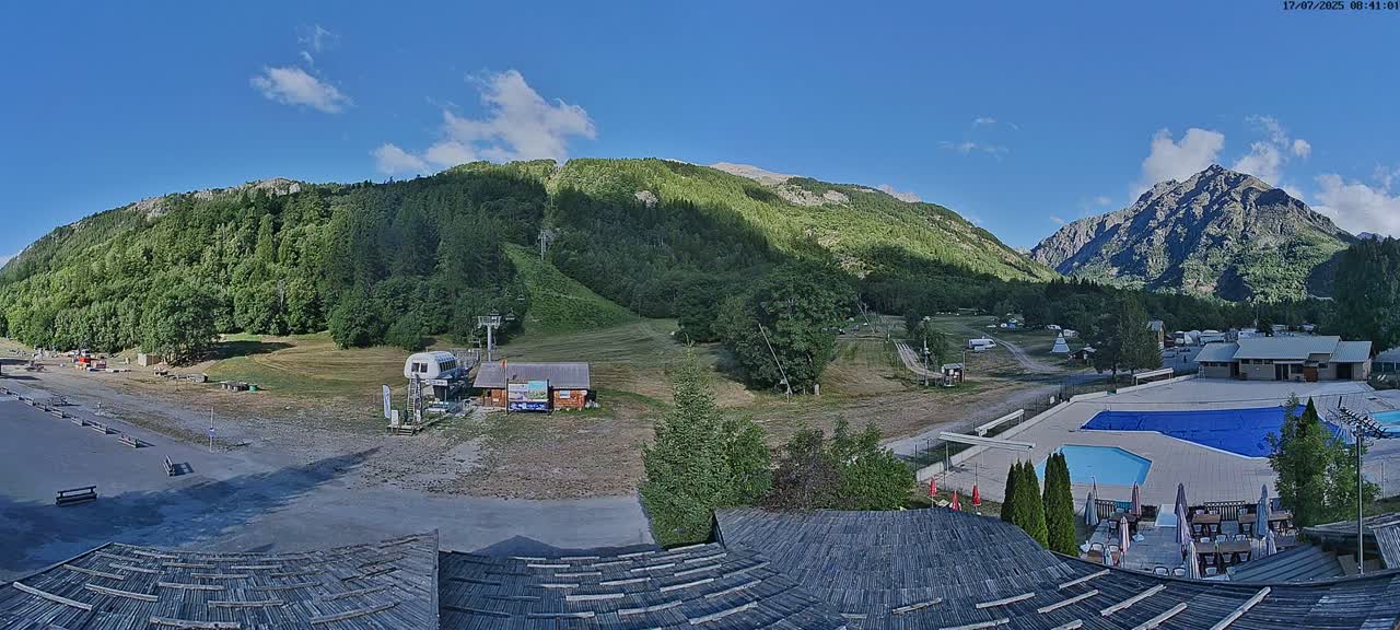 A mountain valley scene on a clear, sunny day shows a ski lift, grassy hills, a swimming pool, and buildings nestled among green trees and mountains.