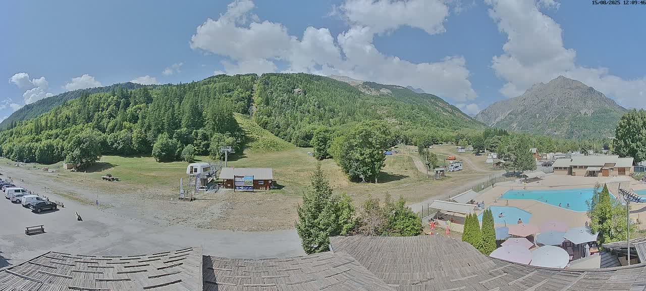 A mountain valley scene on a clear, sunny day shows a ski lift, grassy hills, a swimming pool, and buildings nestled among green trees and mountains.