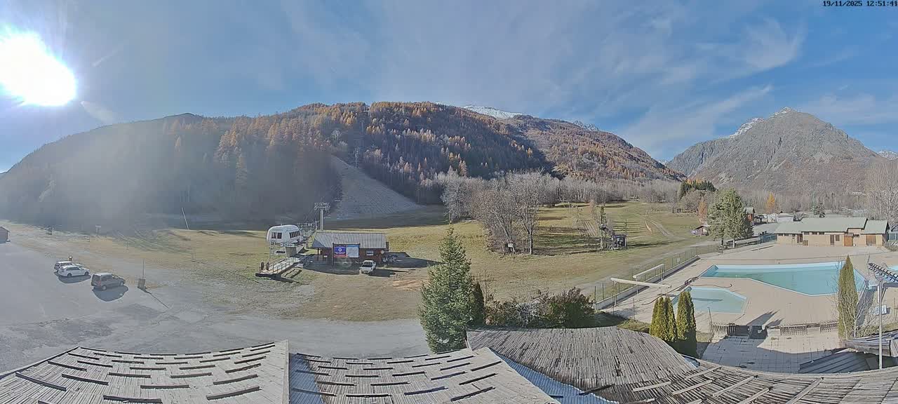 A mountain valley scene on a clear, sunny day shows a ski lift, grassy hills, a swimming pool, and buildings nestled among green trees and mountains.