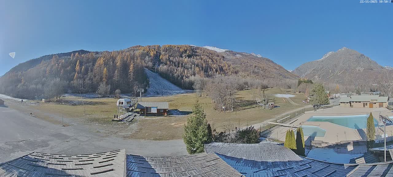 A panoramic view reveals a mountainous landscape featuring a partially snow-dusted ski slope with a lift, small buildings, a swimming pool complex, and trees with autumnal hues or bare branches, all under a clear blue sky on a bright, cold day.