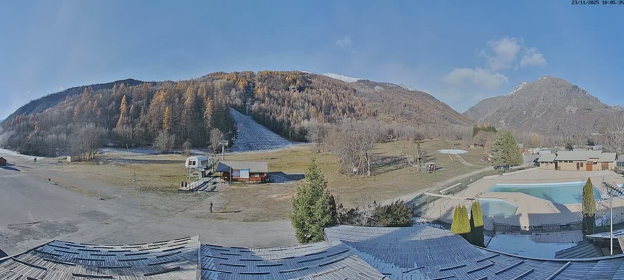 A wide panoramic view captures a mountain valley with forested hillsides, a partially frosted ski slope, and scattered buildings including a ski lift and covered swimming pool, all under a clear, sunny sky with visible patches of frost on the ground and snow on distant peaks, suggesting a crisp late autumn or early winter day.