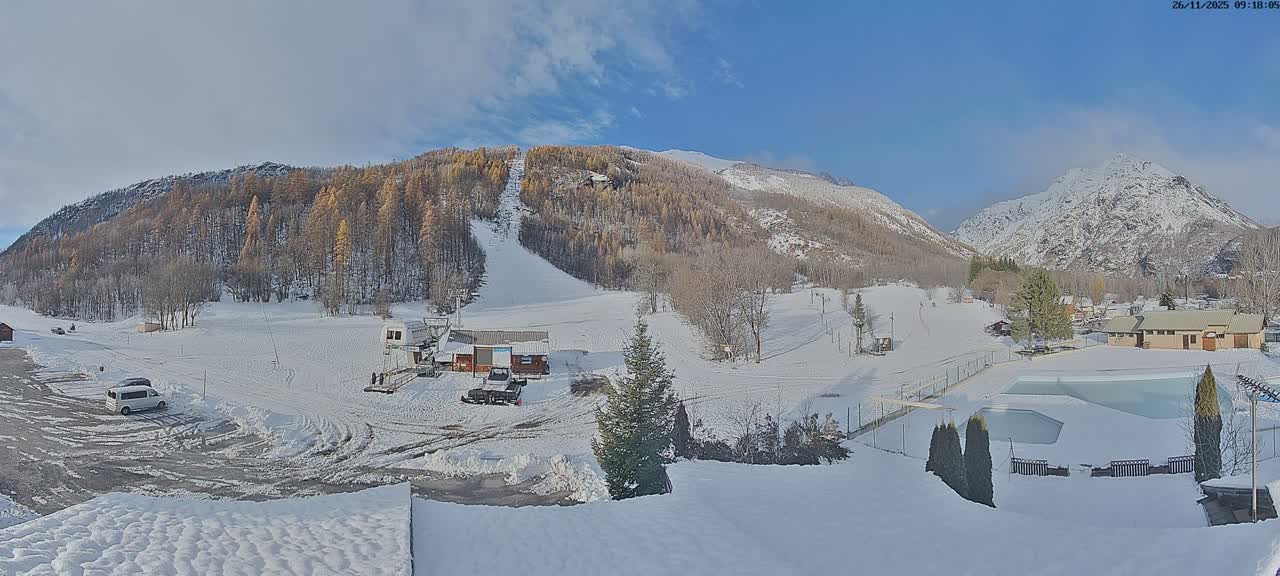 A wide panoramic view reveals a snow-covered mountain valley with a ski slope and lift, golden-leafed and bare trees, a few buildings, a snowy parking lot with vehicles, and a frozen outdoor pool area, all under a clear to partly cloudy blue sky.