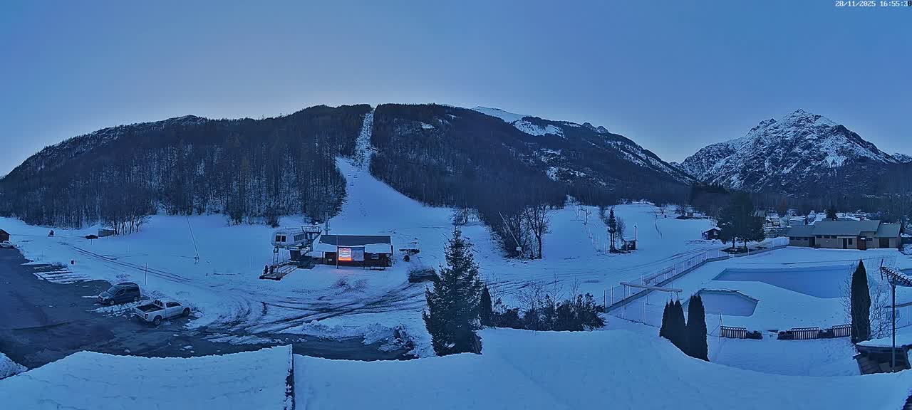 A wide view of a snowy mountain ski resort at dusk under a clear blue sky, featuring an illuminated ski lift base building, several ski slopes, parked vehicles, and a snow-covered outdoor pool complex.