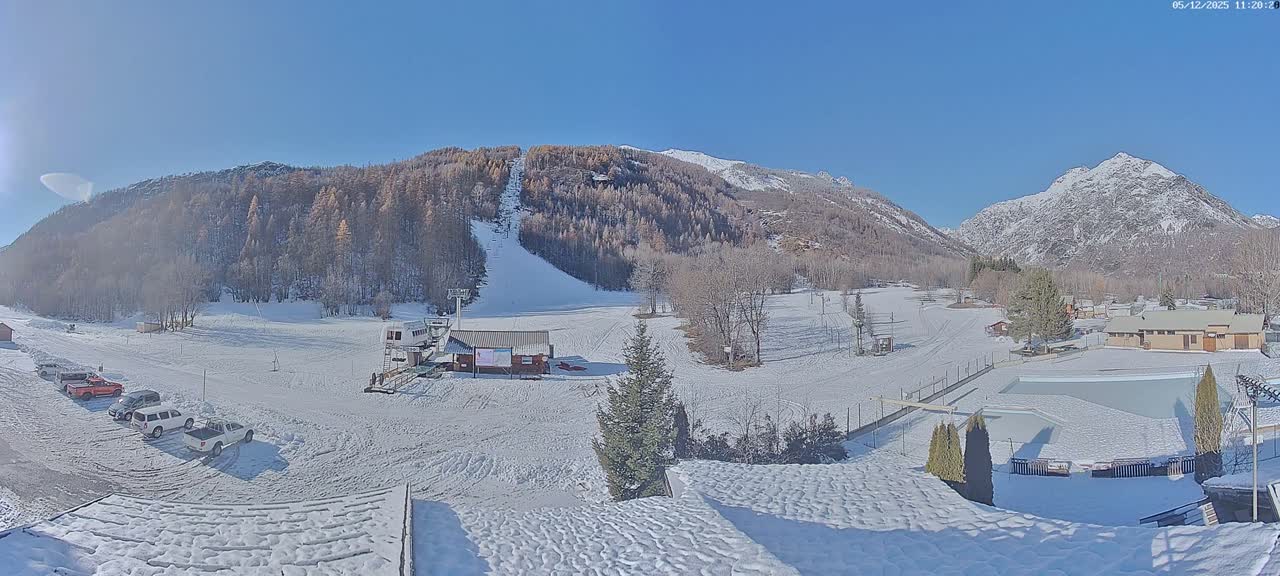 A wide panoramic shot reveals a snow-covered mountain landscape featuring a ski slope with a lift, various buildings, and parked cars, all under a clear blue sky on a sunny winter day.