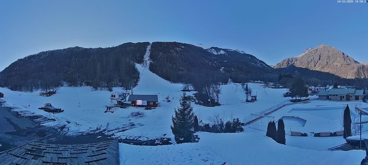 A wide panoramic shot reveals a snow-covered mountain landscape featuring a ski slope with a lift, various buildings, and parked cars, all under a clear blue sky on a sunny winter day.