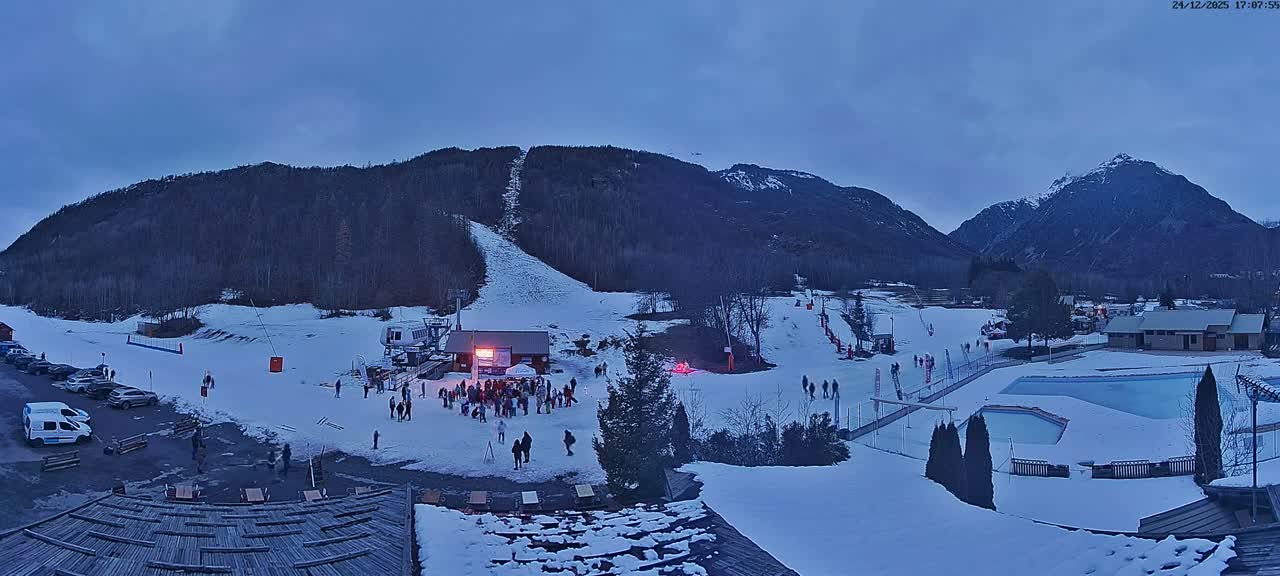 A wide panoramic shot reveals a snow-covered mountain landscape featuring a ski slope with a lift, various buildings, and parked cars, all under a clear blue sky on a sunny winter day.