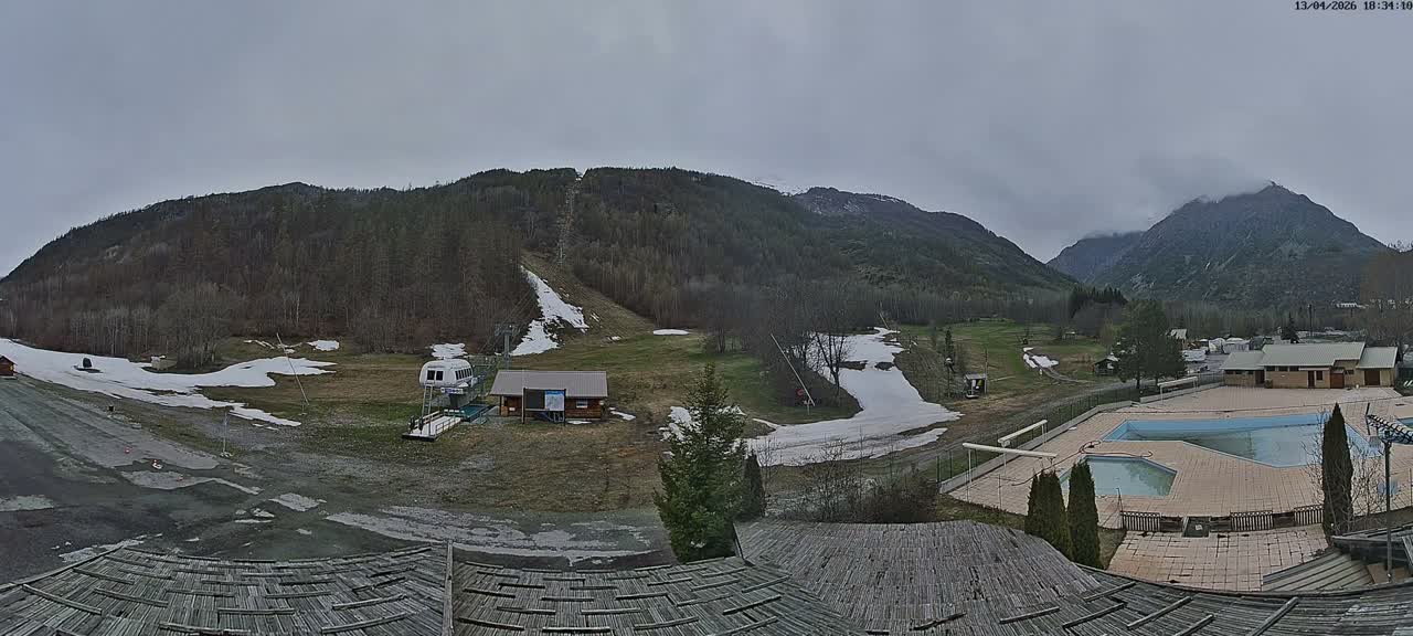 A wide panoramic shot reveals a snow-covered mountain landscape featuring a ski slope with a lift, various buildings, and parked cars, all under a clear blue sky on a sunny winter day.
