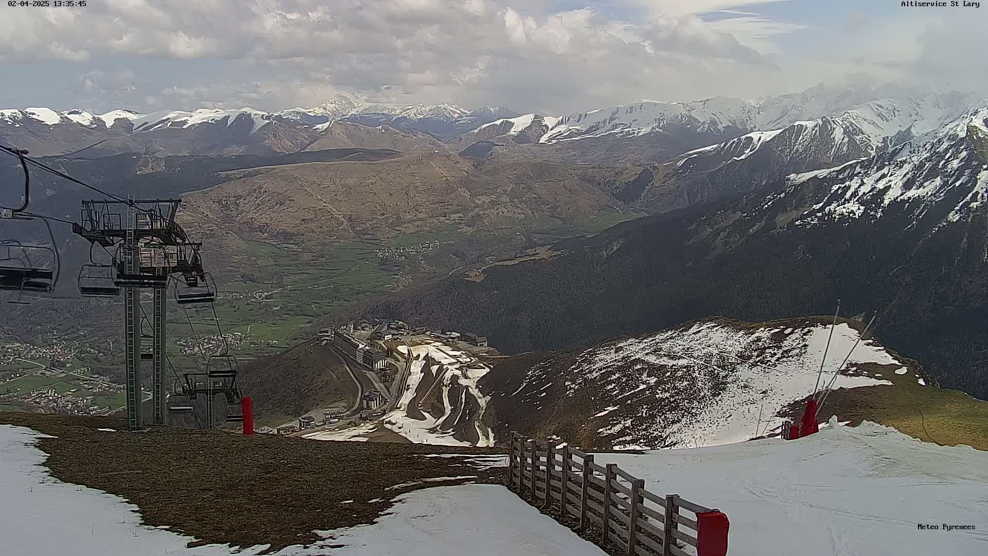 A chairlift stands atop a mountain overlooking a valley with snow-capped mountains in the distance under partly cloudy skies.
