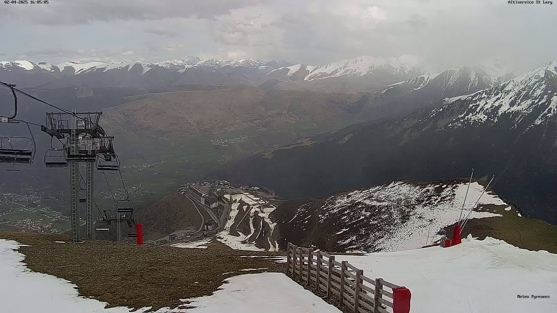 A ski lift overlooks a mountain valley with snow-covered peaks under an overcast sky.