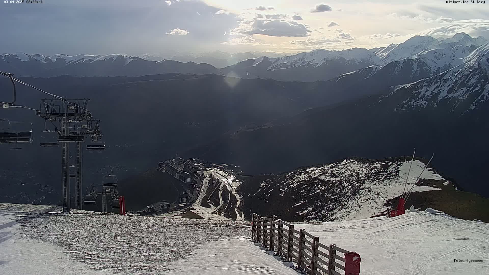 A mountaintop ski resort scene under partly cloudy skies, showing a ski lift, snow-covered slopes, and distant snow-capped mountains.