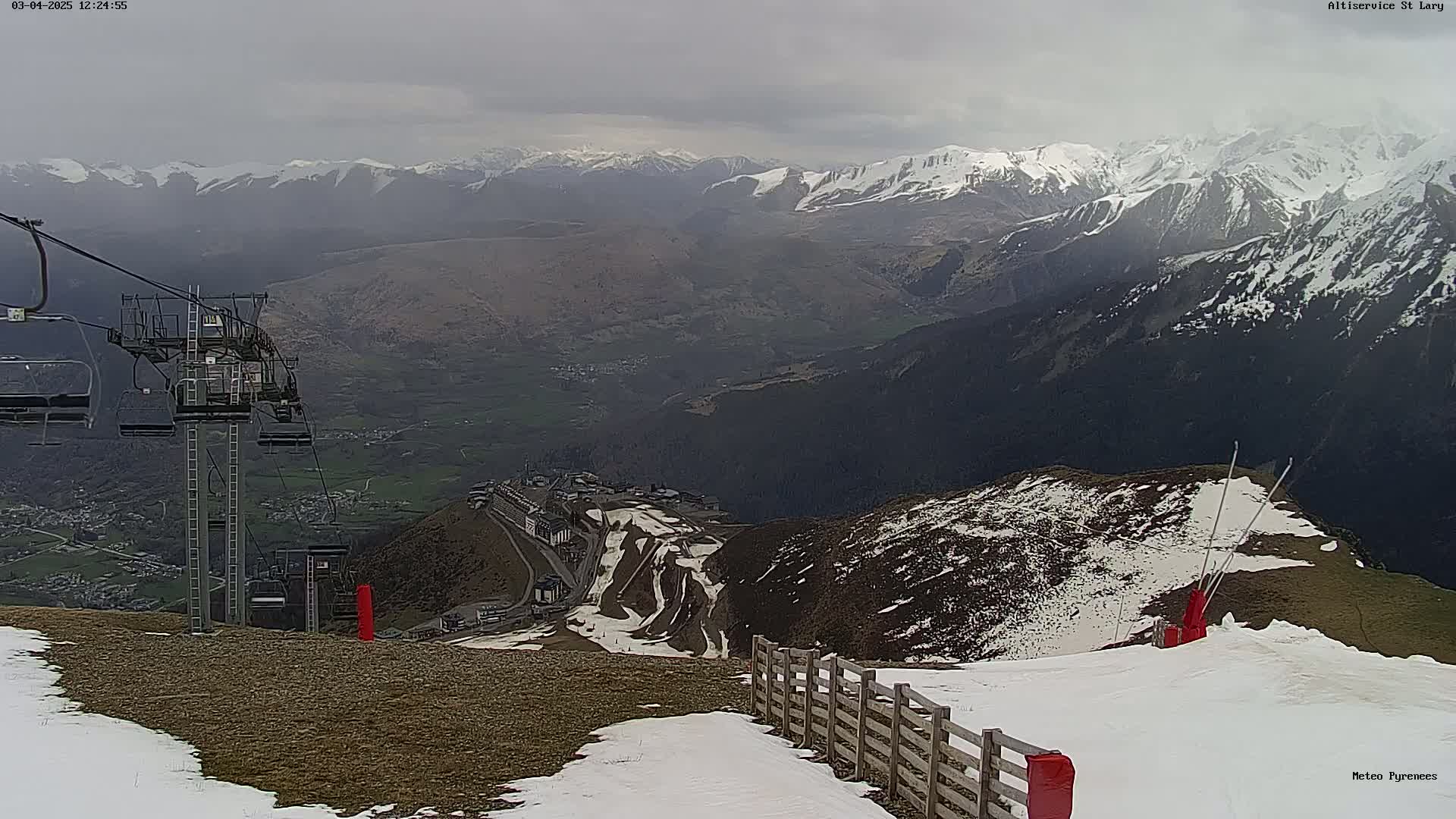 A mountain ski resort on an overcast day, with snow patches on the slopes and snow-capped mountains in the distance.