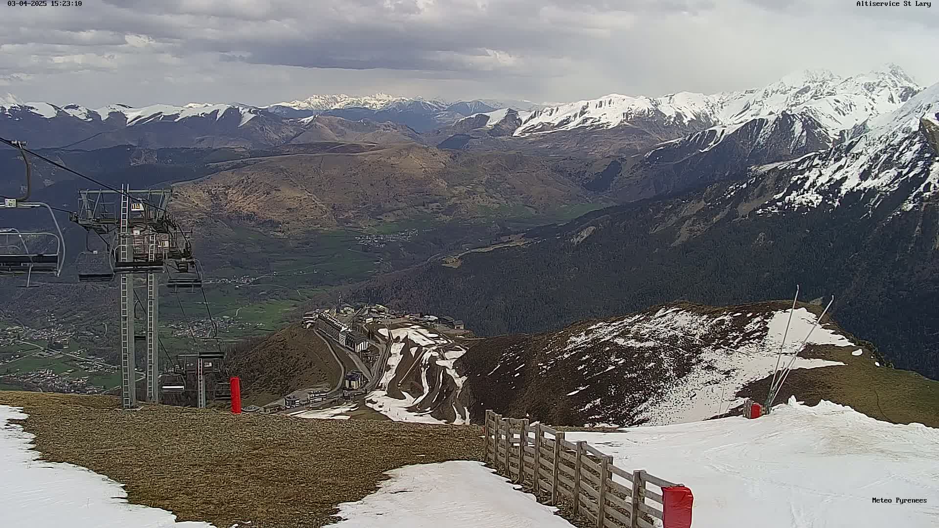 A mountaintop ski lift overlooks a valley with snow-capped mountains in the distance under cloudy skies.