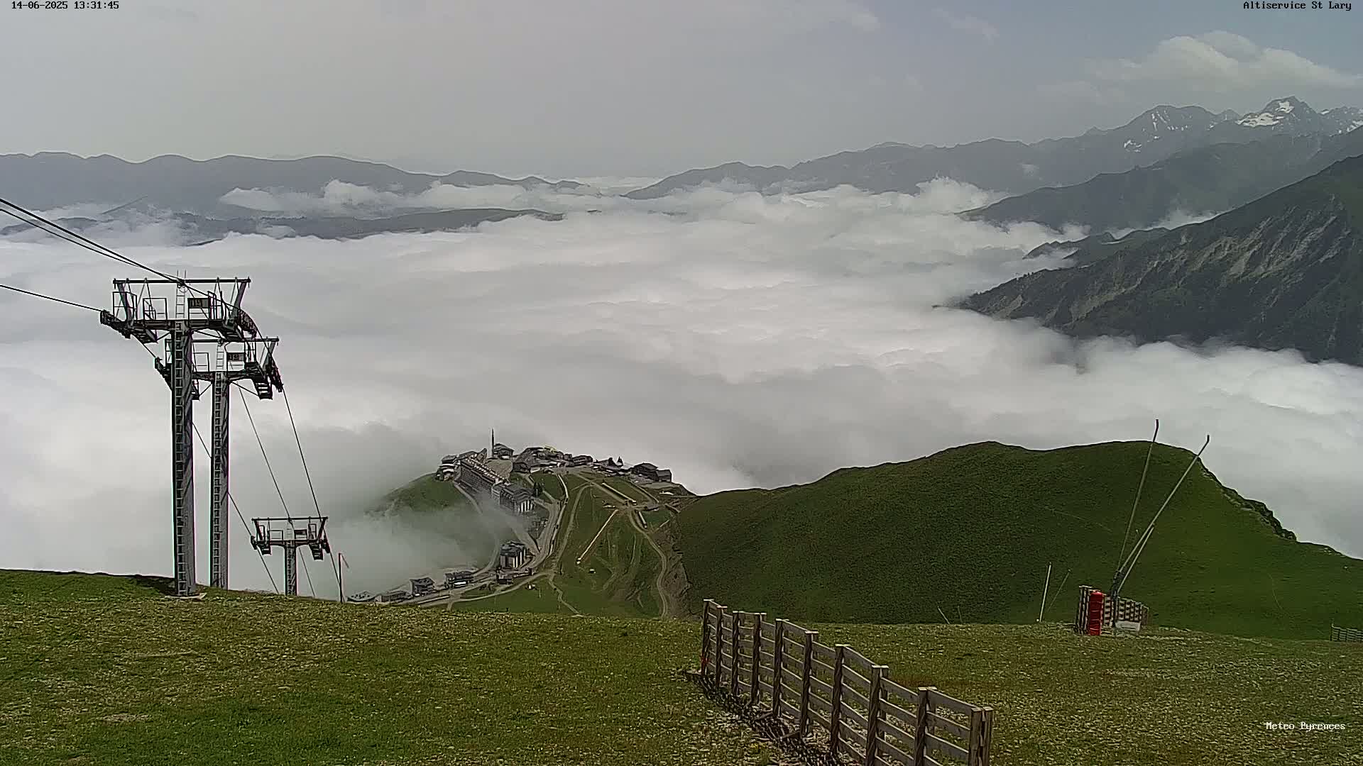 A mountaintop view shows a chairlift and a village nestled in a valley below, all enveloped in a thick layer of fog with distant mountains partially visible above the clouds.