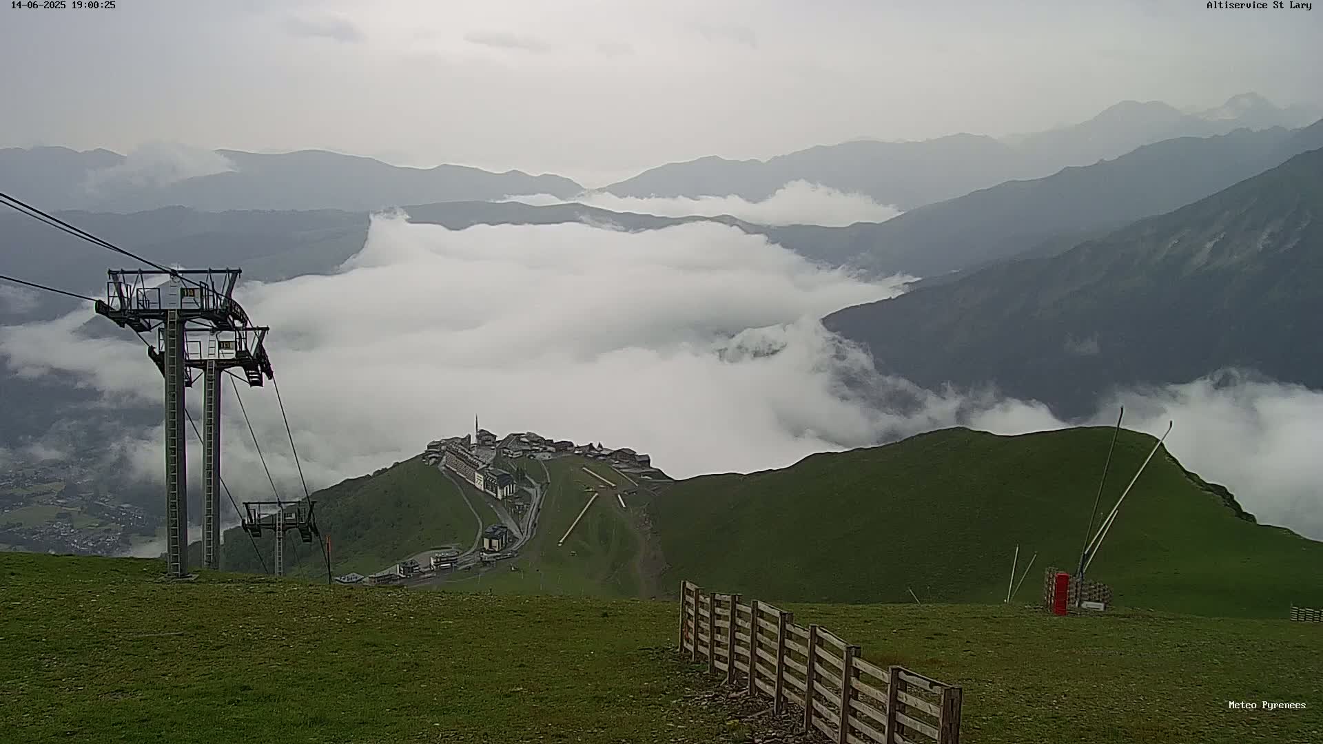 A mountaintop scene shows a chairlift, a wooden fence, and a valley shrouded in low clouds under an overcast sky.