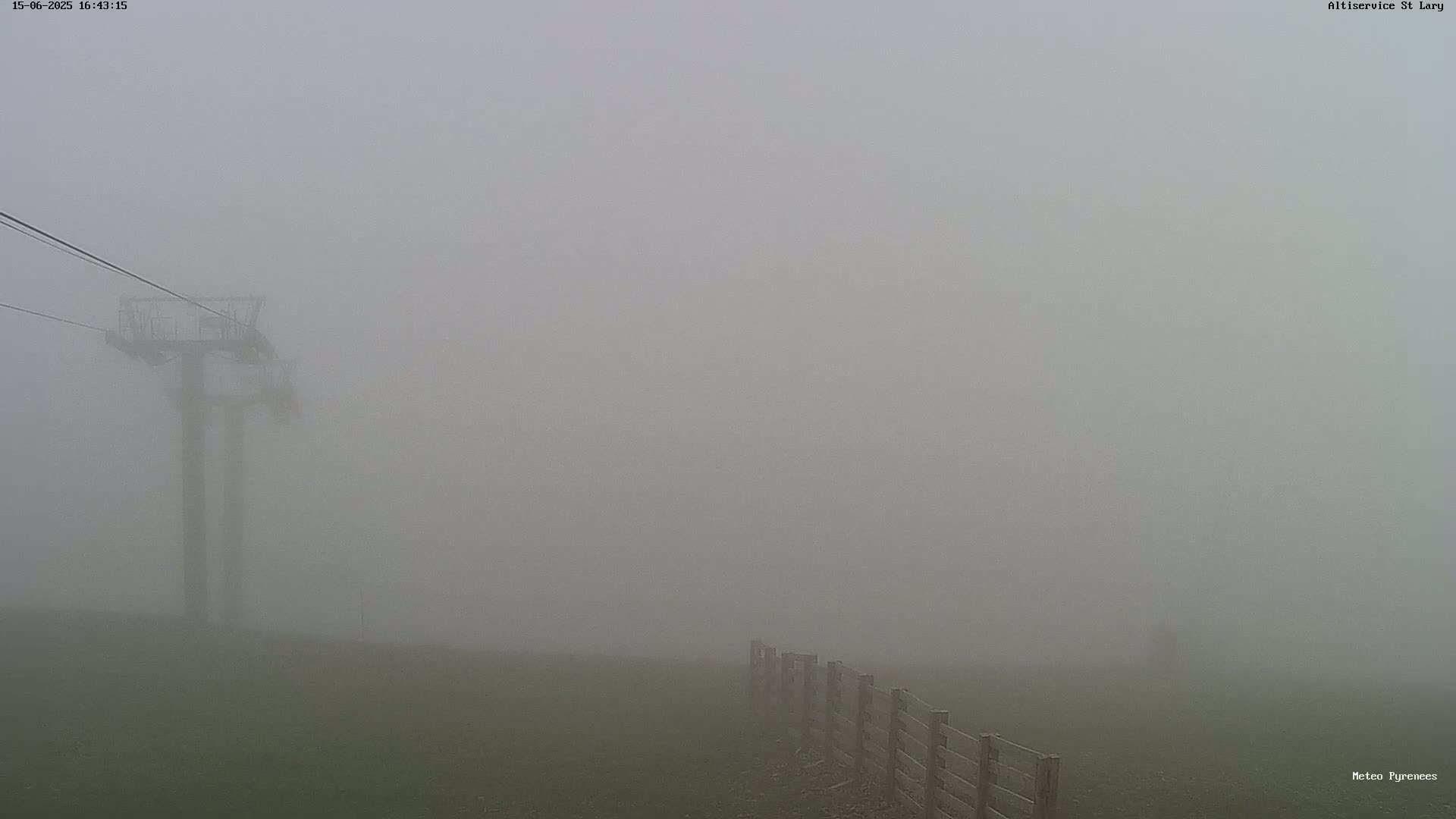 A chairlift stands in a thick fog obscuring a grassy field and wooden fence.