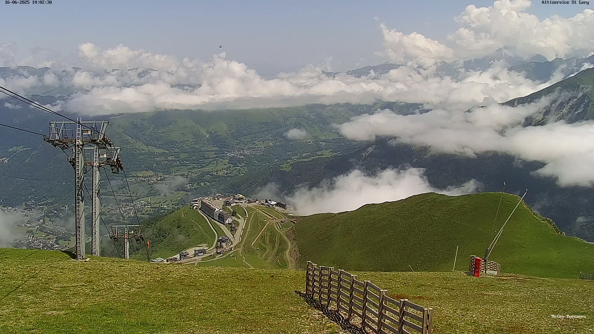A mountaintop view on a partly cloudy day shows a ski resort nestled in a valley below, with chairlifts and a wooden fence in the foreground.