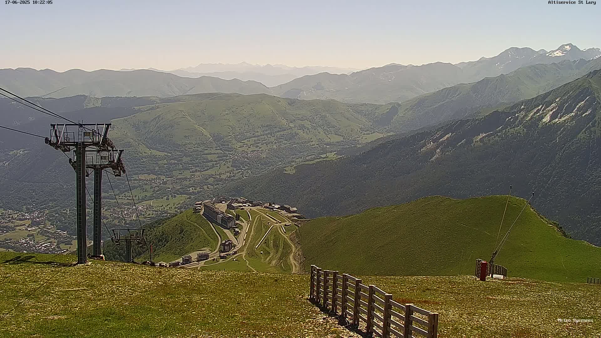 A mountaintop view on a hazy day shows a ski resort nestled in a valley below, with a chairlift and a wooden fence in the foreground.