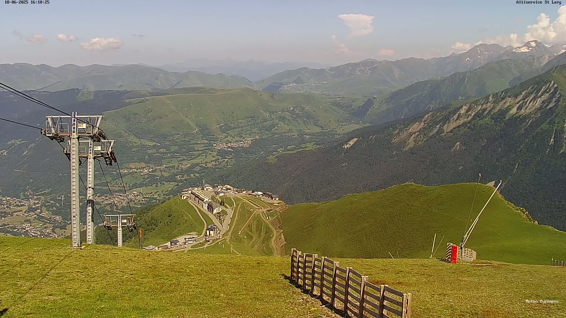A mountaintop view on a partly sunny day shows a valley with a town below, chairlift towers, and a wooden fence in the foreground.