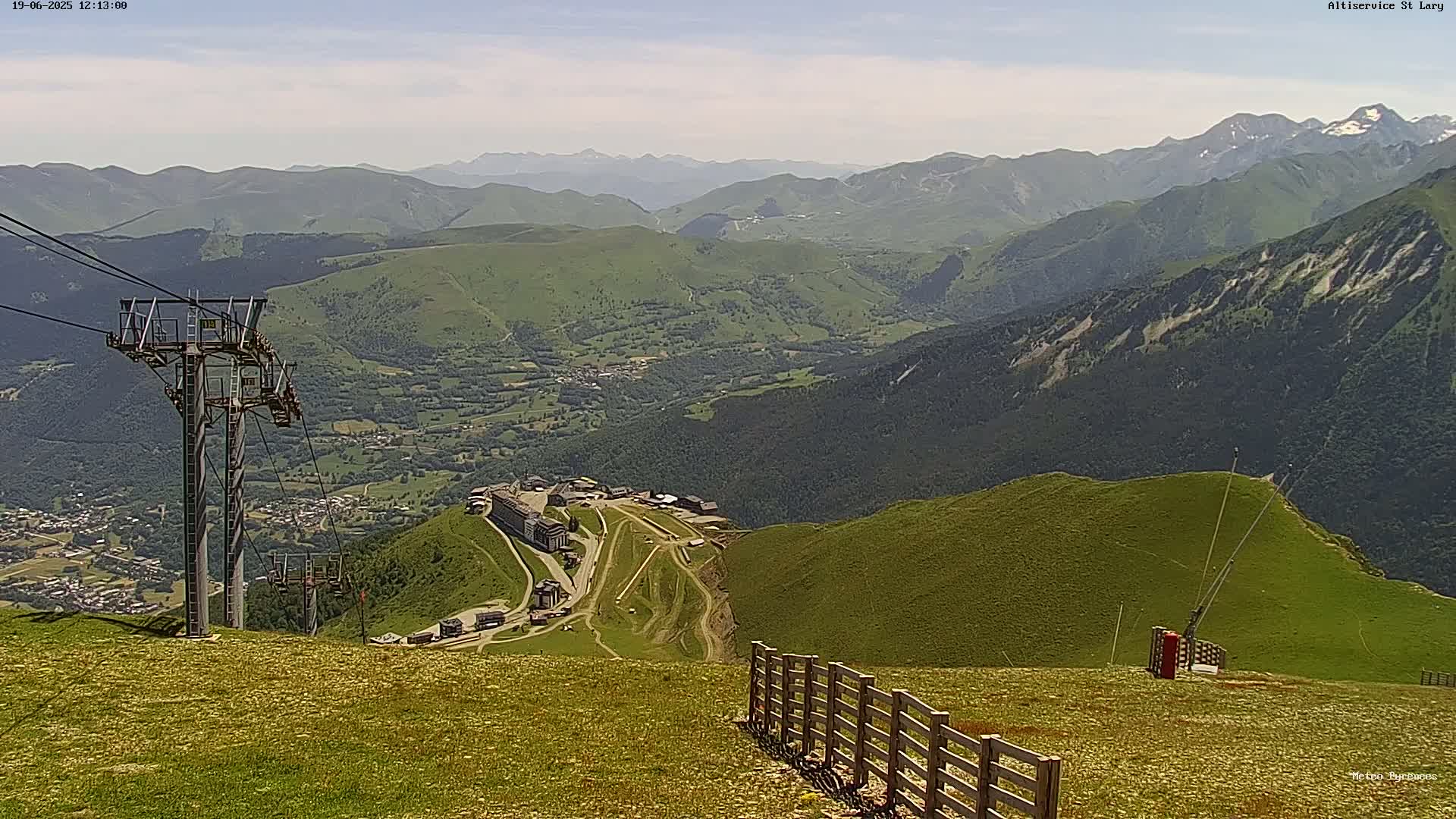 A sunny mountaintop view shows a ski resort nestled in a valley, with a chairlift and fence in the foreground, overlooking a vast expanse of rolling green hills and distant mountains.