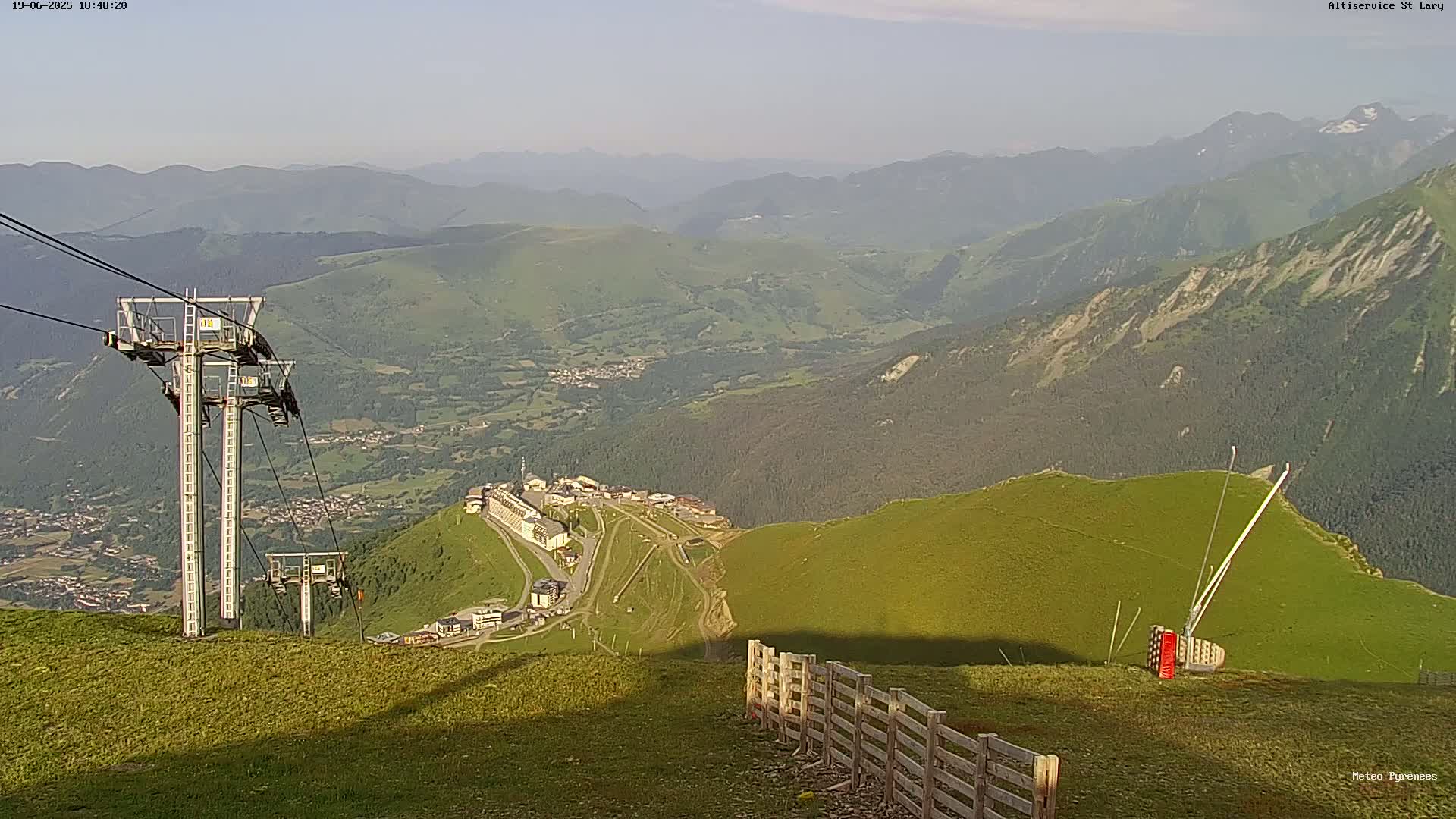 A high-angle, long shot of a mountaintop ski resort with chairlifts, under hazy, sunny conditions, overlooking a valley and distant mountains.