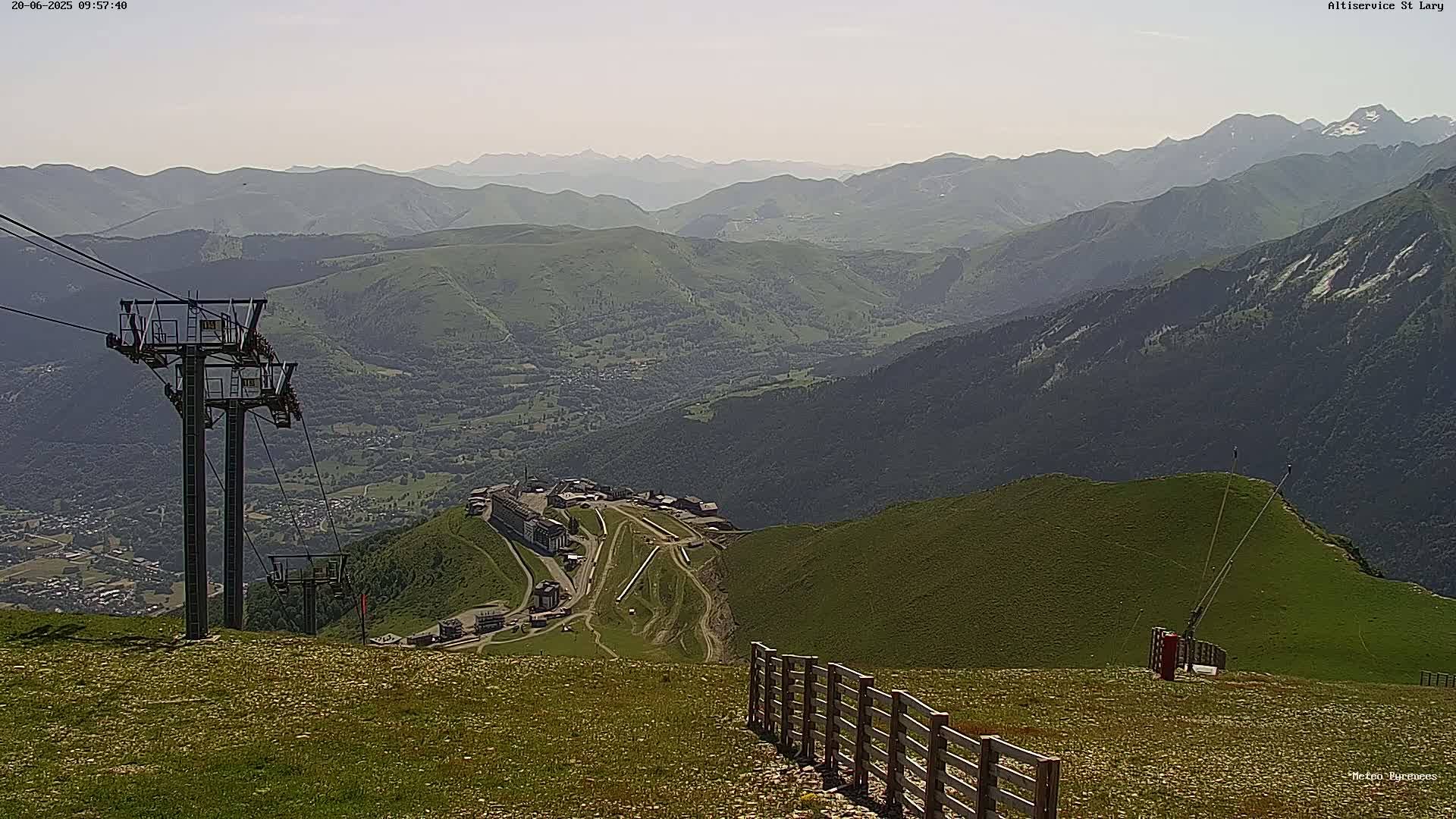 A mountaintop view on a hazy day shows a chairlift, a valley with buildings, and a wooden fence leading down a grassy hillside toward a distant mountain range.
