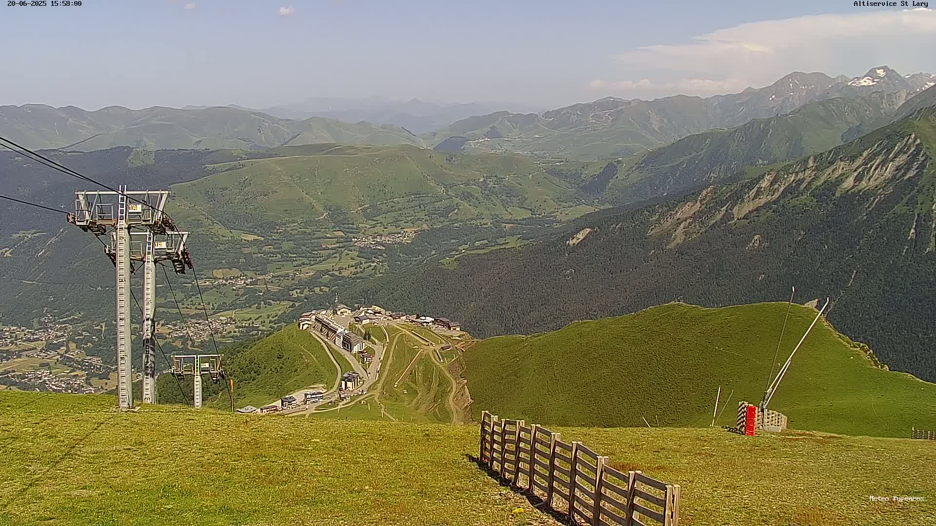 A mountaintop view on a clear day shows a chairlift, a wooden fence, and a valley below with buildings and more mountains in the distance.