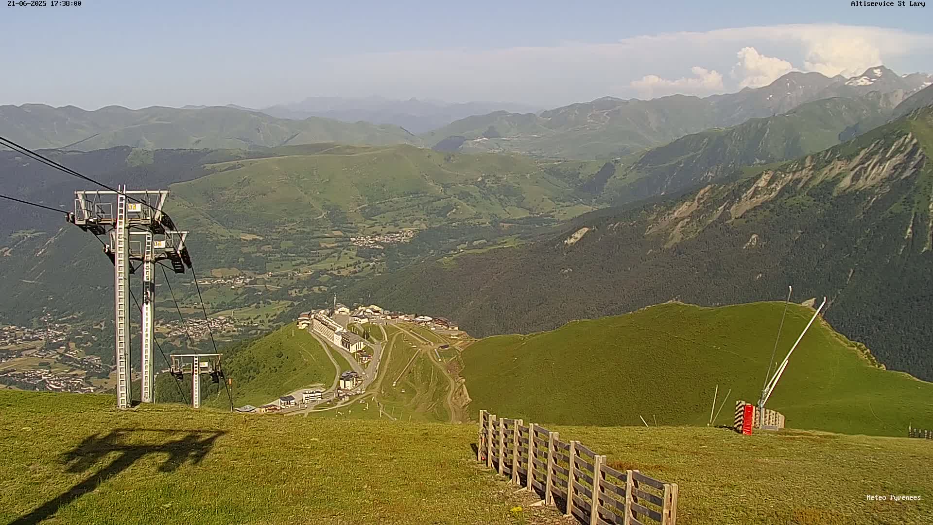 A high-angle view shows a mountainous landscape under partly cloudy skies, with a chairlift and fence in the foreground.
