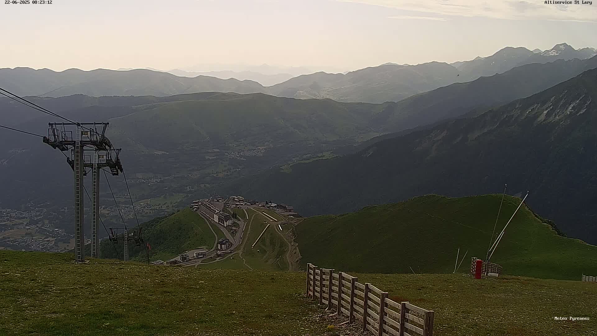 A mountaintop view shows a chairlift, a wooden fence, and hazy mountains under a lightly cloudy sky.